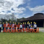 Estudantes posam para foto na área externa da empresa, em um gramado, junto ao um letreiro em tamanho grande que diz "aço é ArcelorMittal". Eles usam capacetes de proteção na cor amarela.