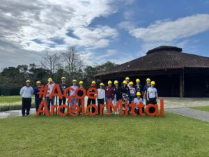 Estudantes posam para foto na área externa da empresa, em um gramado, junto ao um letreiro em tamanho grande que diz "aço é ArcelorMittal". Eles usam capacetes de proteção na cor amarela.