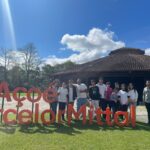 Estudantes posam para foto na área externa da empresa, em um gramado, junto ao um letreiro em tamanho grande que diz "aço é ArcelorMittal".