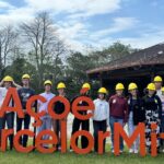 Estudantes posam para foto na área externa da empresa, em um gramado, junto ao um letreiro em tamanho grande que diz "aço é ArcelorMittal". Eles usam capacetes de proteção na cor amarela.