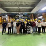 Fotografia dos estudantes e professores, posandopara a foto lado a lado, no interior da empresa. Todos usam capacetes, óculos de proteção e equipamentos de proteção individual.