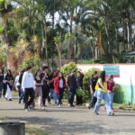 Fotografia na área externa em frente à entrada do setor de avicultura. O grupo de visitantes está caminhando, seguindo a monitora que veste um colete de cor amarela.