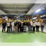 Fotografia dos estudantes e professores, posandopara a foto lado a lado, no interior da empresa. Todos usam capacetes, óculos de proteção e equipamentos de proteção individual.