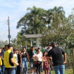 Fotografia na área externa em frente ao laboratório de produção vegetal. Os visitantes observam atentos enquanto o monitor dá explicações sobre a unidade.