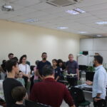Fotografia interna no laboratorio Fábrica de Software. Os visitantes observam atentos enquanto o professor dá explicações sobre o setor.