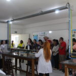 Fotografia na área interna do laboratório de química. Os visitantes observam enquanto a monitora dá explicações sobre o setor. Na imagem é possível verificar as bancadas do labortório e os sistemas de encanamento de gases com diferentes cores.