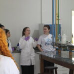 Fotografia na área interna do laboratório de química. A monitora dá explicações sobre o setor aos visitantes. Na imagem é possível verificar as bancadas do labortório e os sistemas de encanamento de gases com diferentes cores.