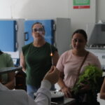 Fotografia interna no laboratório de química. Em primeiro plano, a mão do monitor segura um objeto fino, semelhante a uma caneta. A ponta deste objeto está em chamas e verifica-se um fio de fumaça saindo dali. Em segundo plano, duas mulheres observam atentasà demonstração.