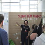 Fotografia na área interna do setor de Suporte e Acompanhamento Educacional. A pedagoga fala sobre o setor aos visitantes, que observam com atenção. Ao fundo, um letreiro em uma parede em que se lê "sejam bem-vindos".
