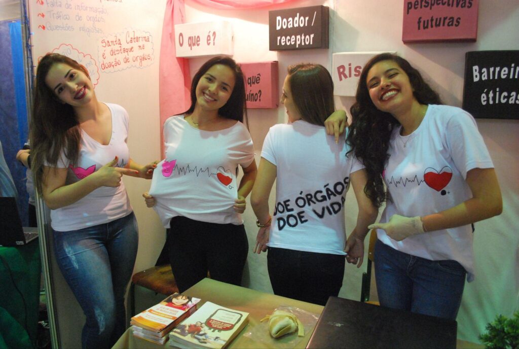 Quatro estudantes posam para foto em um estande. Uma delas veste uma camiseta que diz "doe órgãos". Nas paredes do estande, mensagens relacionadas ao tema.