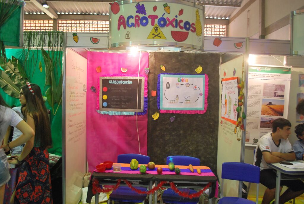 Fotografia do estande "Agrotóxicos". Nas paredes ao fundo, metade da parede está enfeitada com uma faixa vertical na cor rosa e a outra metade com uma faixa vertical na cor preta. Nas duas faixas há cartaes de cartolina com informações. No centro do estande, sobre duas mesas, estão expostos alguns vegetais, como pimentão, tomate, maçã, pepino e cenoura.
