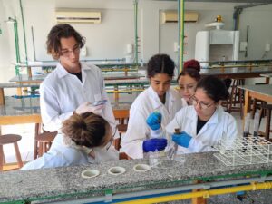 Grupo de alunos realizam um experimento em uma bancada em um laboratório de química. Eles vestem jalecos brancos e luvas azuis.