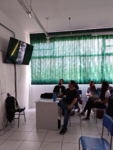 Grupo de estudantes observa imagens em uma TV fixada na parede de uma sala de aula.