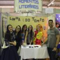 Foto do estande do projto Momentos Literaários. A imagem contém cinco estudantes vestidas de formas diversas, ao lado de um professor. Todos sorriem para a foto. Uma das estudantes estáfantiasiada como a boneca Emília. No fundo do estande, há um letreiro em que se lê "linha do tempo".