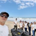 Fotografia de um professsor e um grupo de estudantes. Eles estão em uma praia, de costas para o mar. Em frente deles há alguns sacos cheio de lixo.