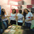 Quatro estudantes posam para foto em um estande. Uma delas veste uma camiseta que diz "doe órgãos". Nas paredes do estande, mensagens relacionadas ao tema.