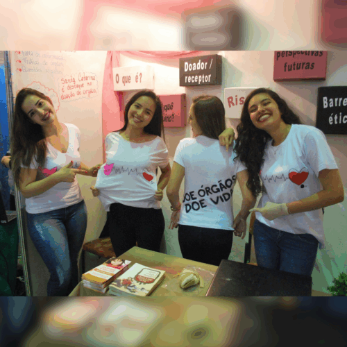 Quatro estudantes posam para foto em um estande. Uma delas veste uma camiseta que diz "doe órgãos". Nas paredes do estande, mensagens relacionadas ao tema.