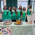 Grupo Além do Sabor. Meninas com avental verda. Em frente a elas, uma mensa com alimentos coloridos.