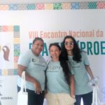 Fotografia de três estudantes da EJA-EPT do IFC Araquari, sendo duas mulheres e um homem, em frente ao banner do evento. À esquera da imagem, uma mulher negra xn cabelo curto; ao centro, uma mulher branca com cabelo liso e longo; à direita, um homem com óculos escursos e cabelos pretos e longos. Os três usam a camiseta do evento, de cor azul clara.