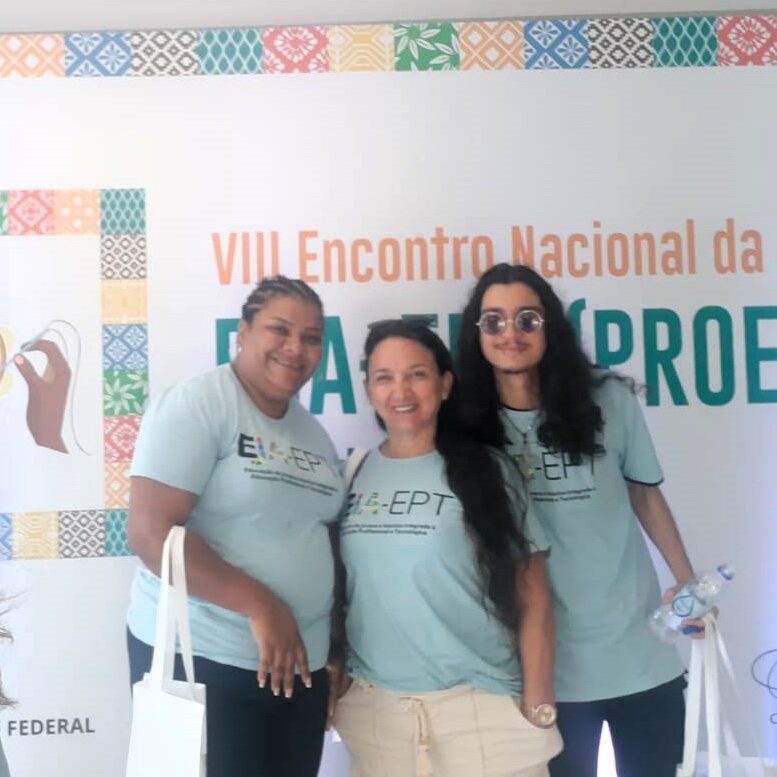 Fotografia de três estudantes da EJA-EPT do IFC Araquari, sendo duas mulheres e um homem, em frente ao banner do evento. À esquera da imagem, uma mulher negra xn cabelo curto; ao centro, uma mulher branca com cabelo liso e longo; à direita, um homem com óculos escursos e cabelos pretos e longos. Os três usam a camiseta do evento, de cor azul clara.